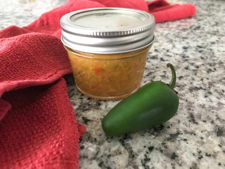 Small jar of peach pepper jam sits on a granite countertop with a jalapeno in the foreground and a red dish towel to the side