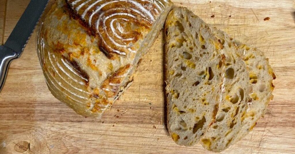 Image of a freshly baked loaf of cheddar garlic herb sourdough bread sitting on a wood cutting. Three pieces have been sliced and lie next to the loaf. In the top left corner is a bread knife.