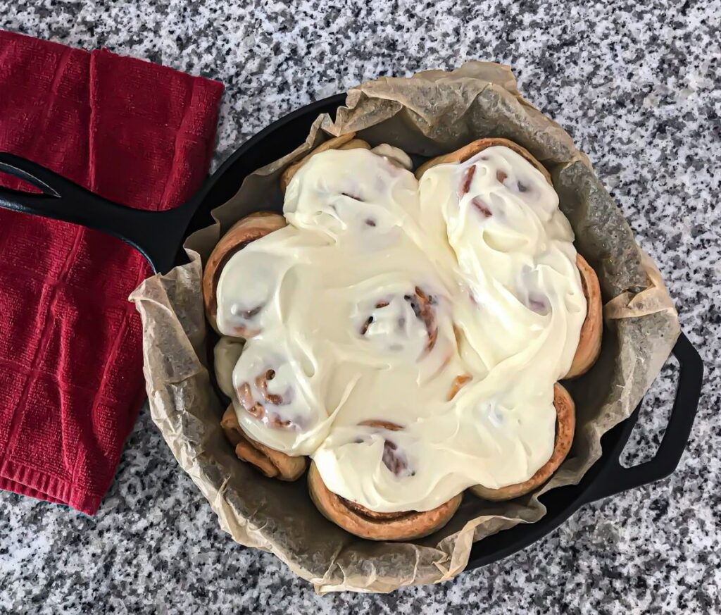 Image of iced sourdough cinnamon rolls in a cast iron skillet sitting on a granite countertop with a red dish towel laid to the side.