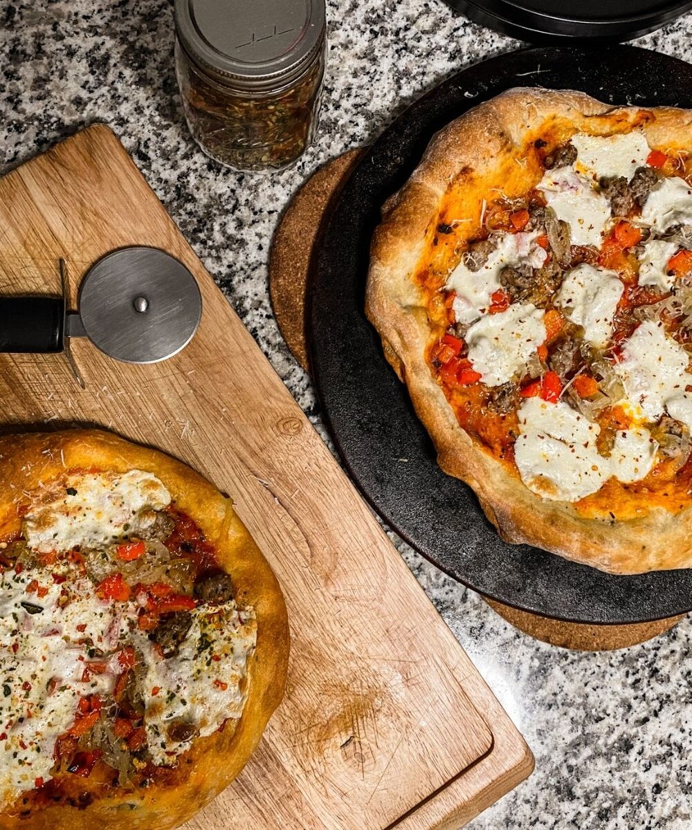 Image of two sourdough pizzas, the larger on the right sits on a pizza stone while the smaller on the left sits on a wooden cutting board.