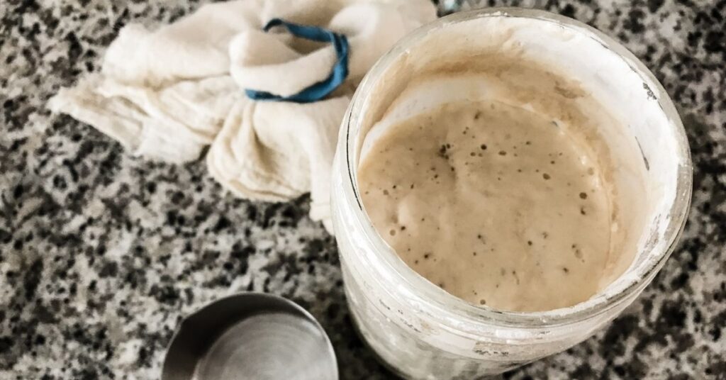 Image of a bubbly, ready to bake with sourdough starter in a jar sitting on a granite countertop. To the left is a metal measuring cup and the cheesecloth cover for the jar.