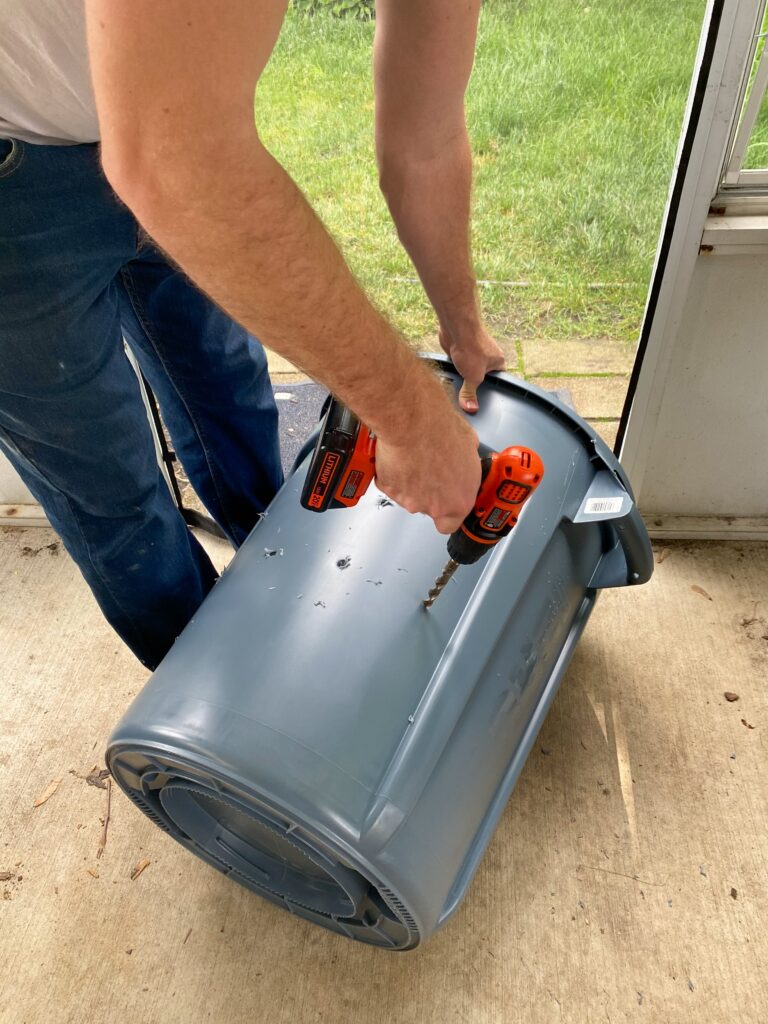 Image of the compost bin in progress. A man's arm drills holes in the side of a plastic outdoor trash can.