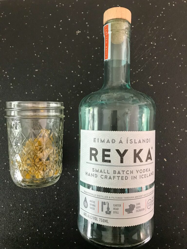 Image of a jar of dried chamomile next to a bottle of Reyka vodka on a black background
