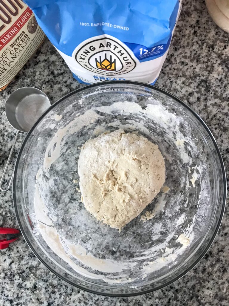 Glass bowl containing autolyse dough with a King Arthur bread flour bag and measuring cup on the counter next to the bowl.
