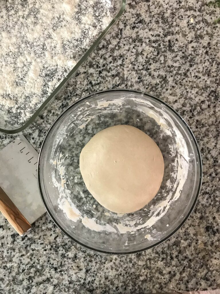 Overhead shot of sourdough pizza crust dough in a glass bowl after being pulled from the fridge the next day. Next to the bowl is a metal dough scraper and a floured glass pan ready to receive the shaped dough balls.