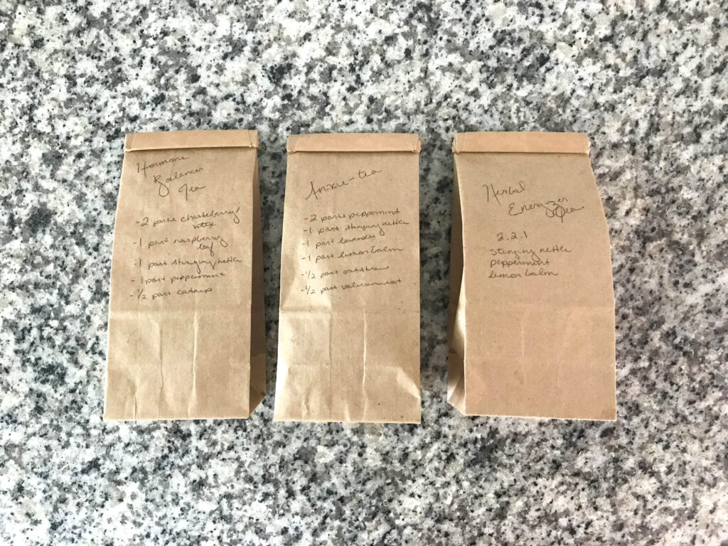 Overhead shot of three brown paper tea bags with blend names and formulations written on each in black pen – herbal tea recipes