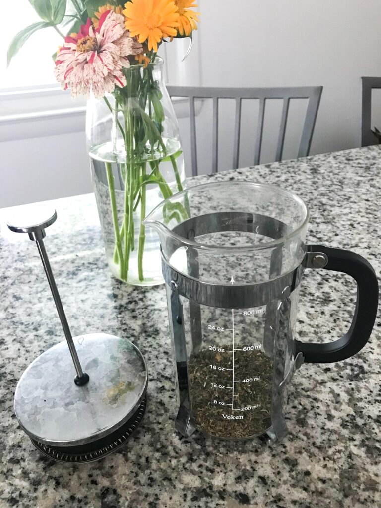 An unfilled French press with herbal tea in the bottom sits on a granite countertop next to its lid. Behind is a glass vase full of zinnias – herbal tea recipes