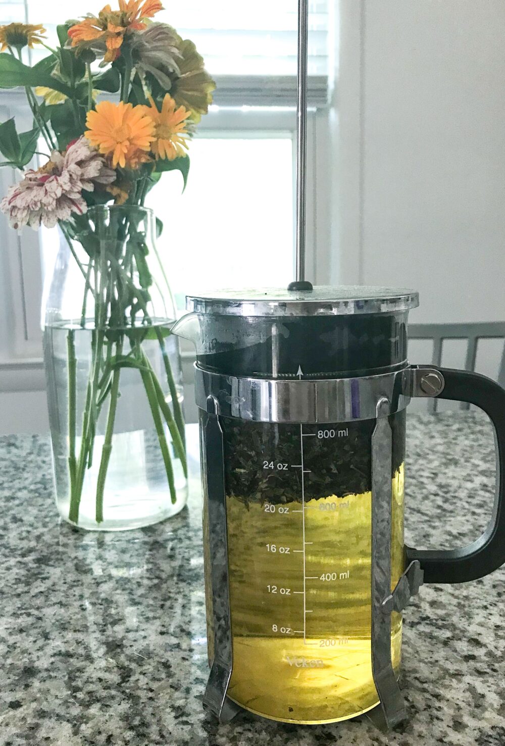 A French pressed filled with steeping herbal tea sits on a granite countertop in front of a bouquet of zinnias in a glass vase – herbal tea recipes