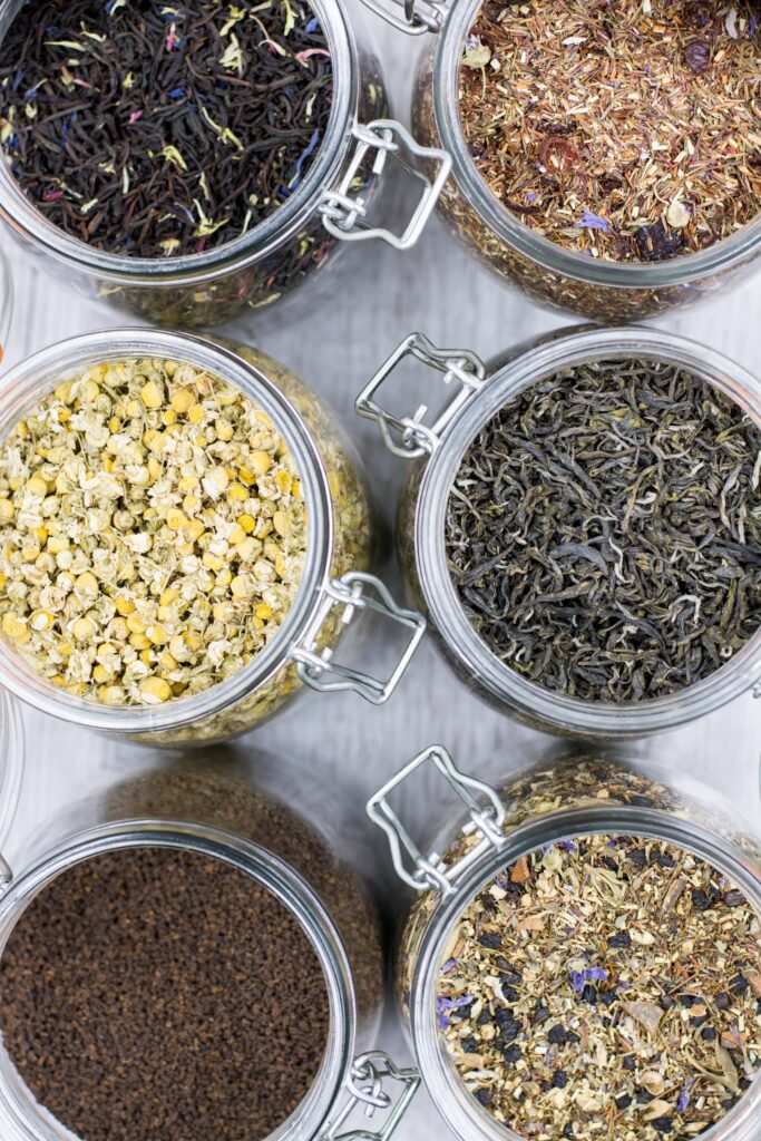 Overhead shot of six open glass swing-top jars, each full of a different dried tea herb – herbal tea recipes