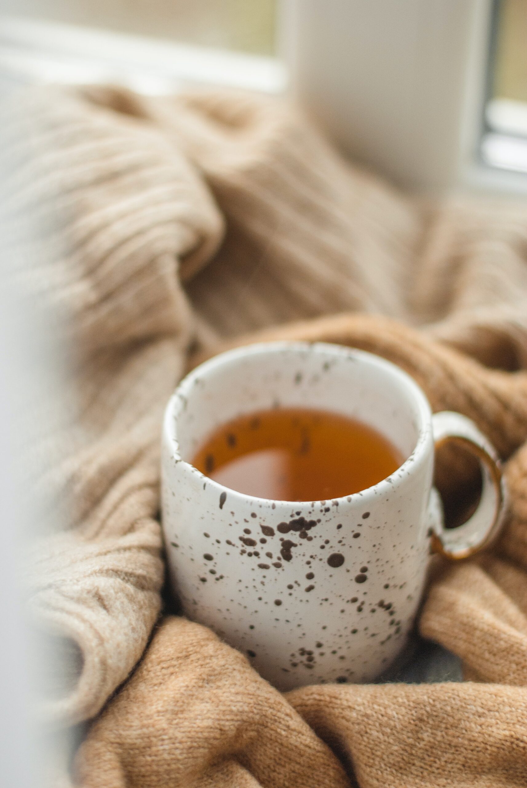 A white and black speckled mug of tea sits on a windowsill with a tan throw blanket – herbal tea recipes