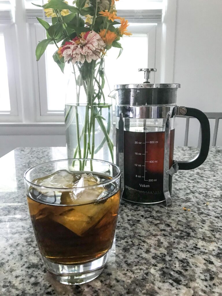 A glass of iced herbal tea sits in front of a full French press of steeped tea and a glass vase containing a zinnia bouquet, on a granite countertop – herbal tea recipes