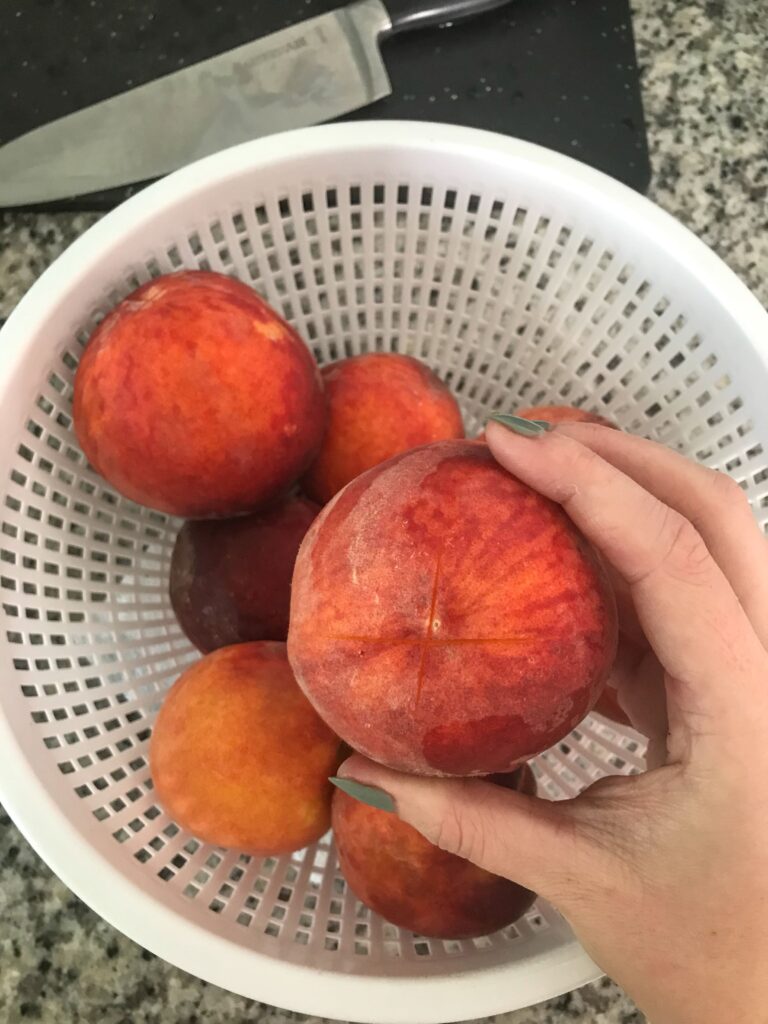 Hand holds a peach above a white colander full of peaches, demonstrating how to cut an X on the bottom before blanching – peach pepper jam