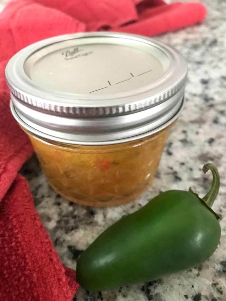 Angle shot of a small jar of peach pepper jam with a jalapeno in the foreground and a red dish towel behind