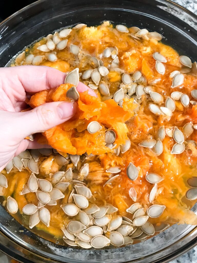 A hand separates pumpkin seeds from guts over a bowl