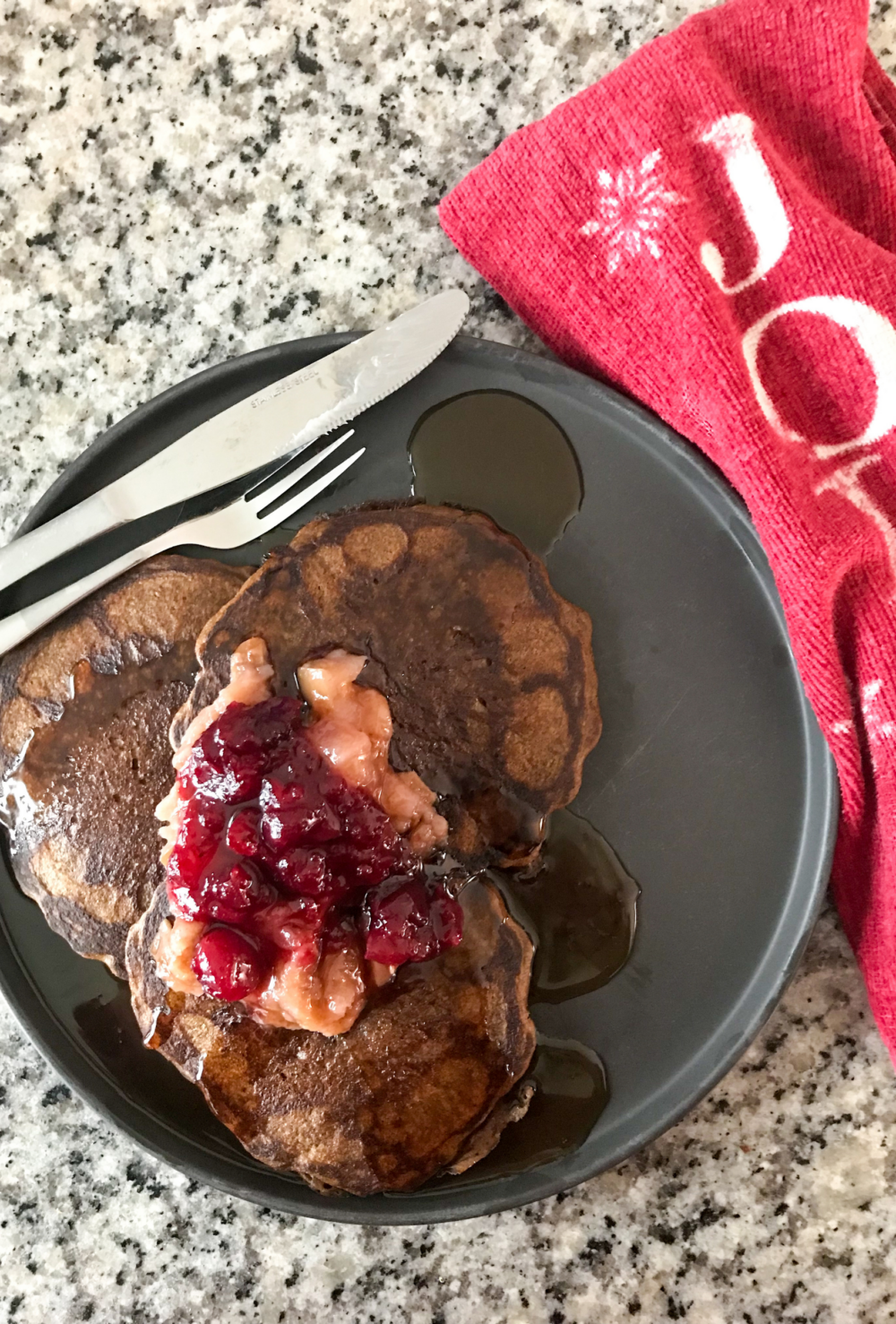 Zoomed out overhead shot of gingerbread sourdough pancakes on a black plate. The pancakes are topped with applesauce, cranberry sauce, and maple syrup. Beside the plate is a red tea towel with the word "jolly" stamped on it.