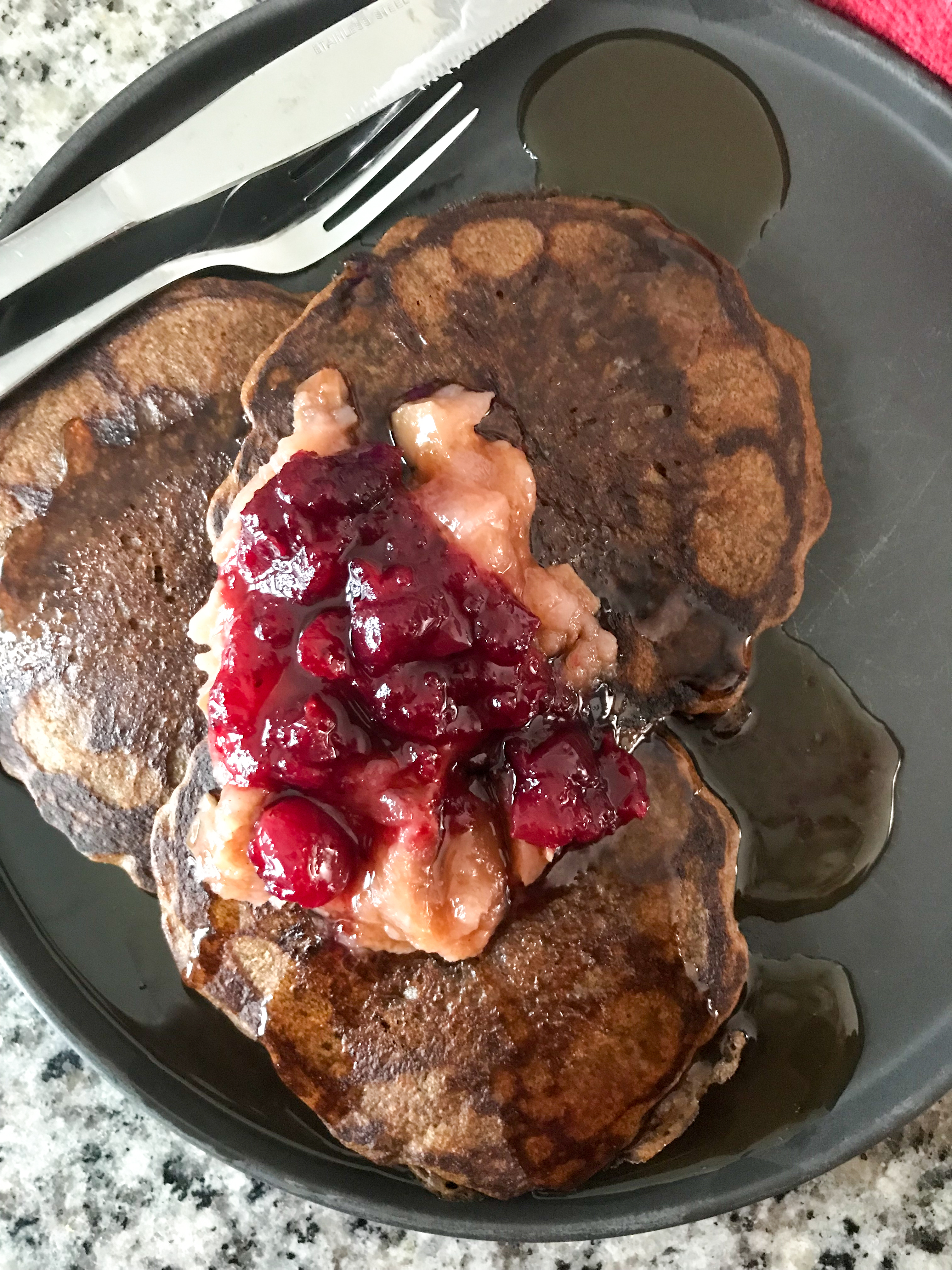 Overhead shot of gingerbread sourdough pancakes on a black plate. The pancakes are topped with applesauce, cranberry sauce, and maple syrup.