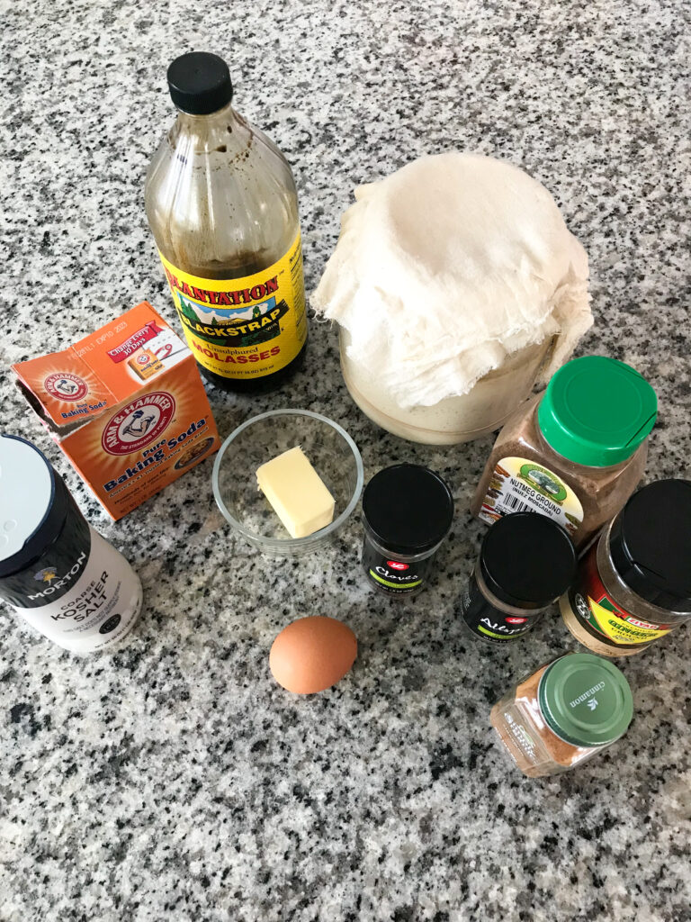 Ingredients for gingerbread sourdough pancakes laid out on a granite countertop. A jar of sourdough starter, baking soda, salt, butter, an egg, and a variety of spices.