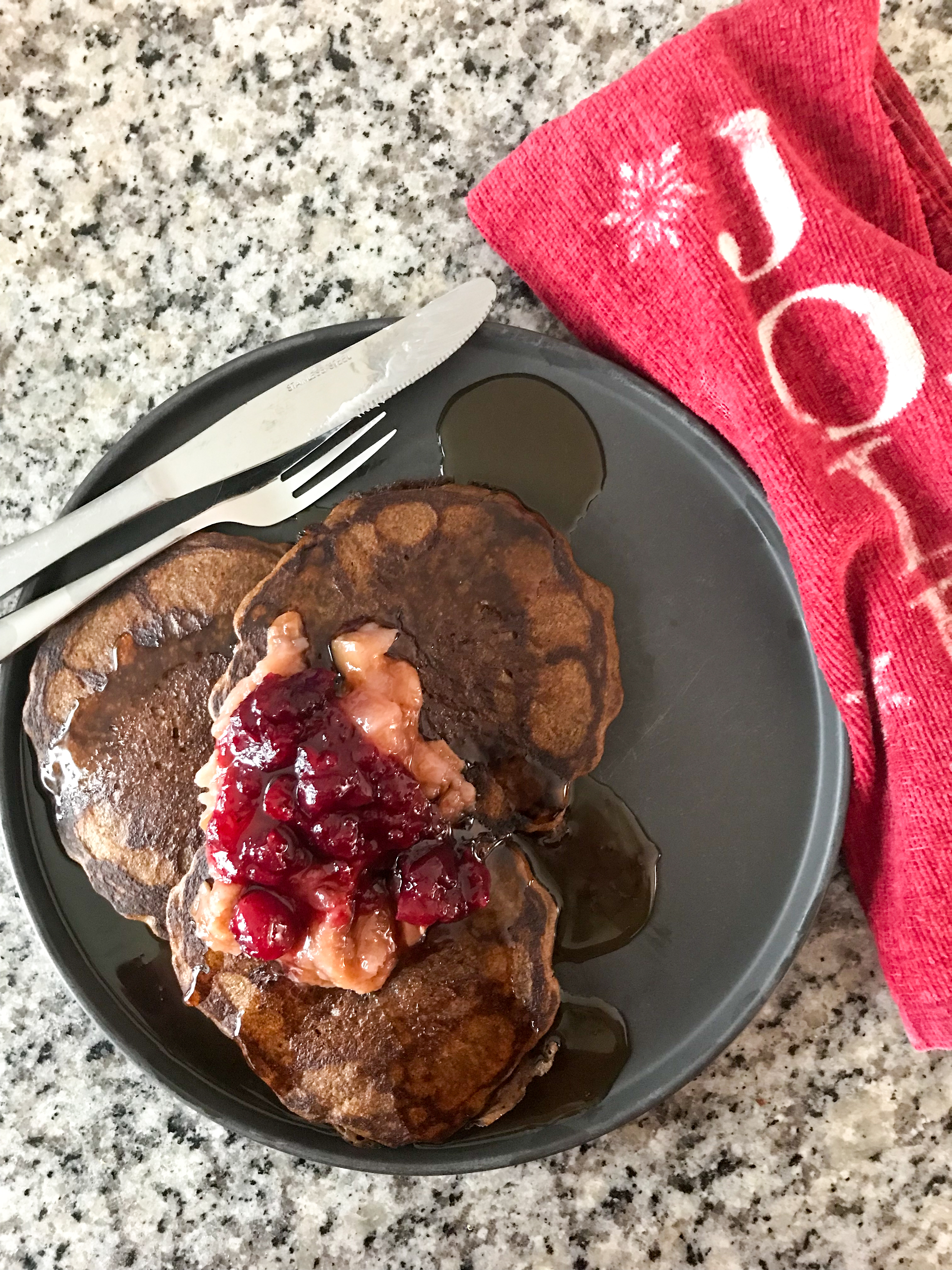 Zoomed out overhead shot of gingerbread sourdough pancakes on a black plate. The pancakes are topped with applesauce, cranberry sauce, and maple syrup. Beside the plate is a red tea towel with the word "jolly" stamped on it.