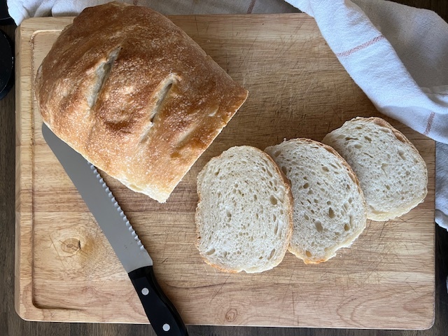 Overhead shot of freshly baked sourdough sandwich bread on a wooden cutting board. Three slices from been cut from the loaf and are lying face-up on the board, as is the serrated bread knife.